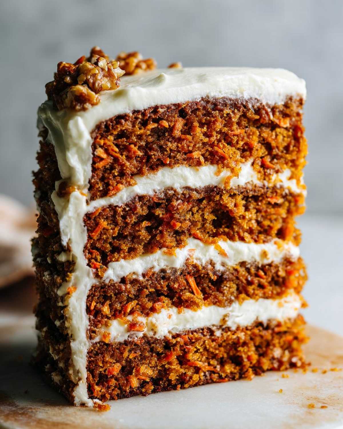 A tall, moist slice of layered carrot cake recipe showing three layers of spiced cake and cream cheese frosting, topped with walnuts.