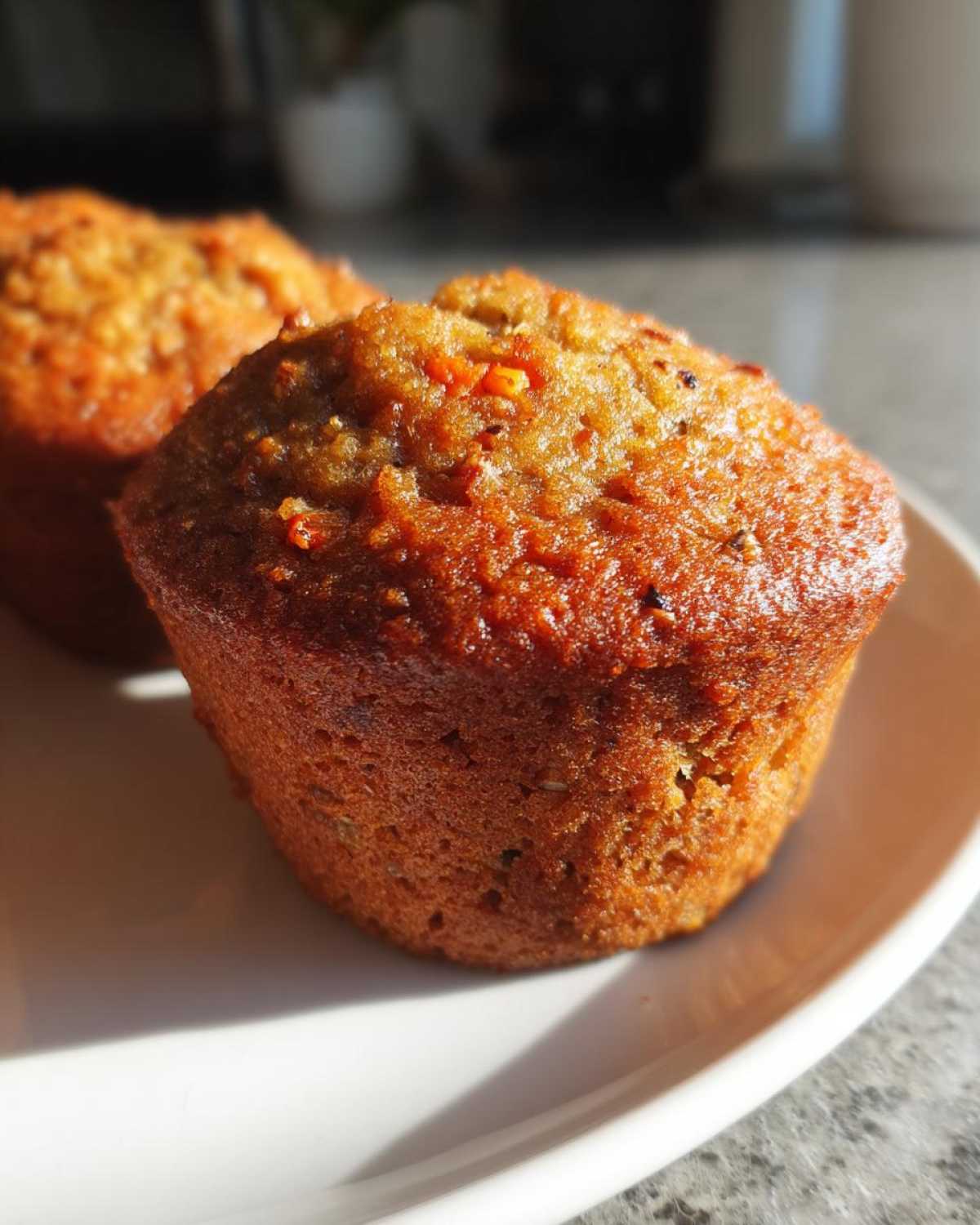 Close-up of a golden brown, moist morning glory muffin sitting on a white plate in bright sunlight.