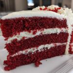 Close-up of a moist slice of red velvet cake showing three layers of deep red cake separated by thick white cream cheese frosting.