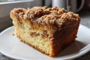 A close-up of a moist slice of sour cream coffee cake featuring a thick cinnamon streusel topping on a white plate.