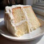 A thick, moist slice of vanilla cake recipe layered with creamy white frosting, sitting on a white plate.