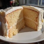 Close-up of a moist vanilla cake recipe slice cut in half, showing two layers and thick white frosting.