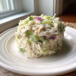 A perfectly molded serving of creamy crab salad mixed with visible chunks of celery and red onion on a white plate.