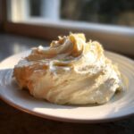 A mound of fluffy, pale yellow cream cheese frosting swirled high on a white plate, backlit by a window.