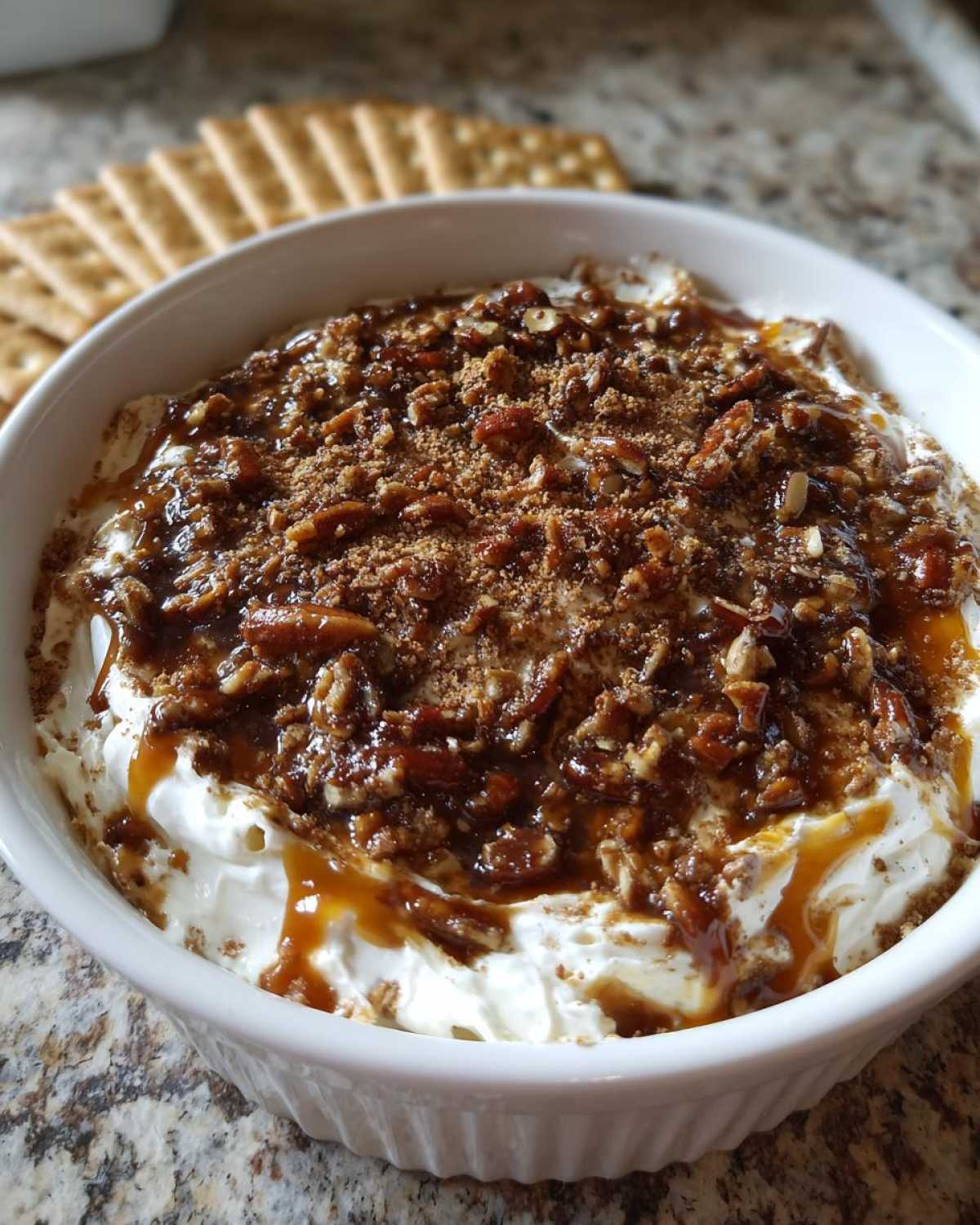 Close-up of a creamy no-bake pecan pie dip topped with pecans and caramel sauce, served with crackers.