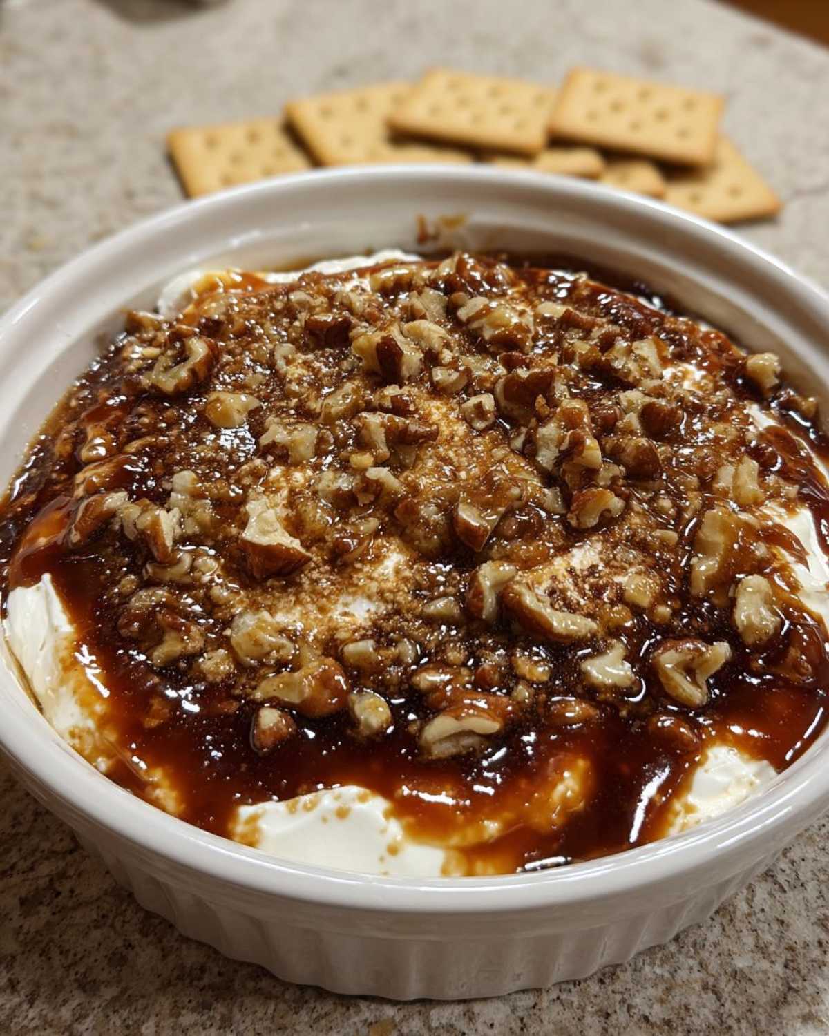 Close-up of a creamy pecan pie dip topped with caramel sauce and chopped pecans, served with crackers.