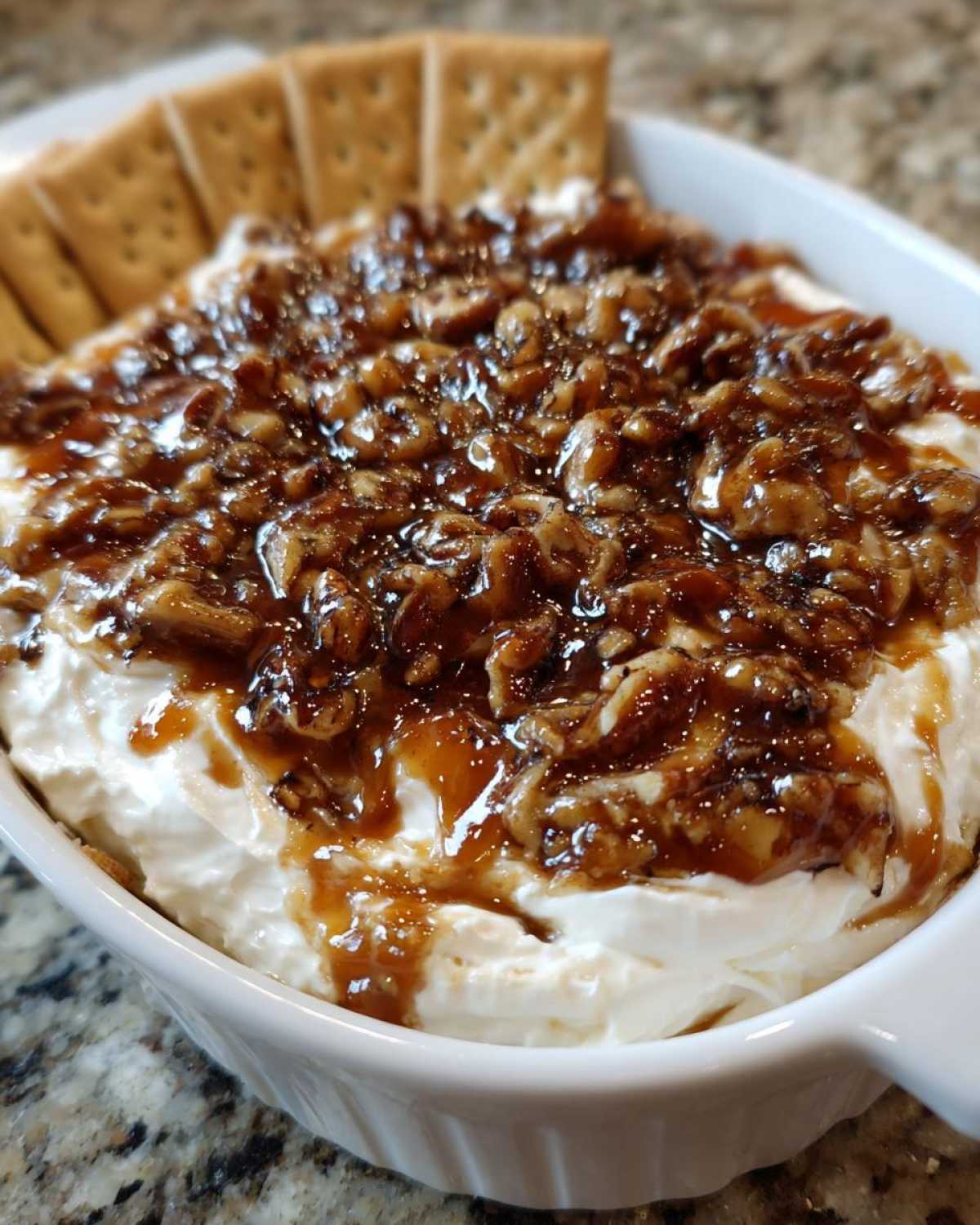 Close-up of a creamy no-bake pecan pie dip topped with candied pecans and caramel sauce, served with crackers.