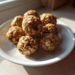 A small pile of homemade, oat-covered protein balls resting on a white plate near a window.
