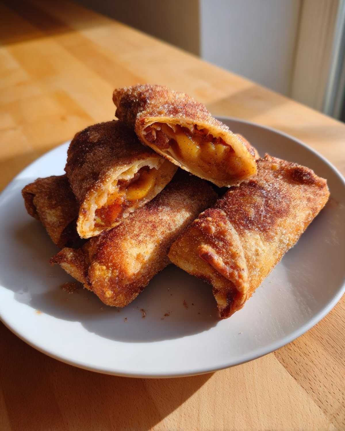A stack of golden brown peach cobbler egg rolls dusted heavily with cinnamon sugar, one is cut open showing the filling.