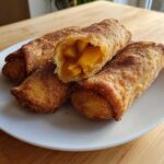Four crispy, golden-brown peach cobbler egg rolls dusted with cinnamon sugar, one cut open to show the warm peach filling.