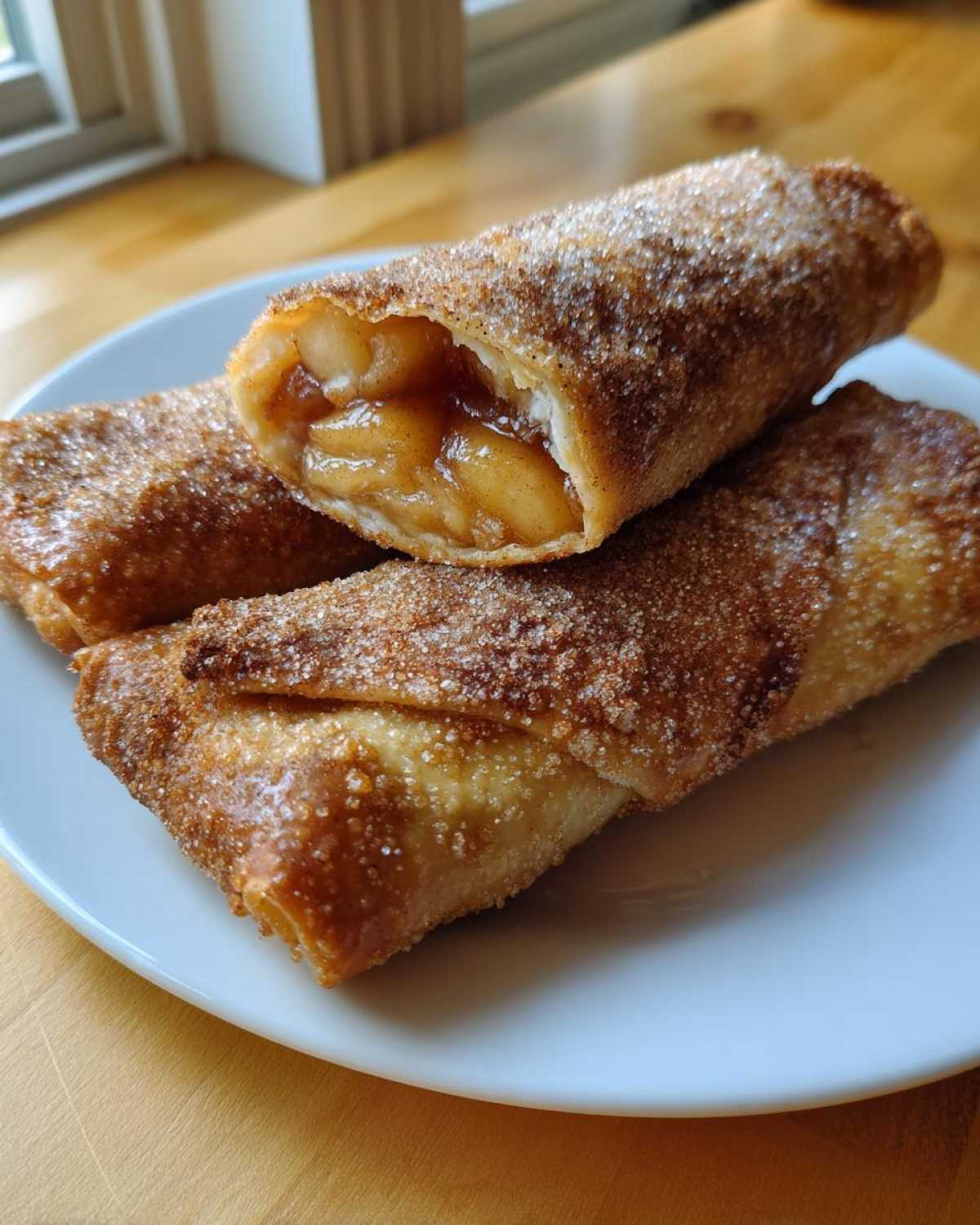 Three crispy, golden-brown peach cobbler egg rolls dusted heavily with cinnamon sugar, one cut open to show the filling.