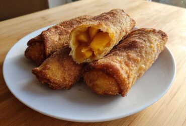 Four crispy, golden-brown peach cobbler egg rolls dusted with cinnamon sugar, one cut open to show the warm peach filling.