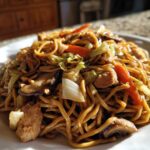 A close-up of a plate piled high with homemade chicken chow mein recipe, featuring noodles, chicken pieces, carrots, and mushrooms.