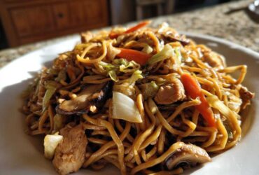 A close-up of a plate piled high with homemade chicken chow mein recipe, featuring noodles, chicken pieces, carrots, and mushrooms.