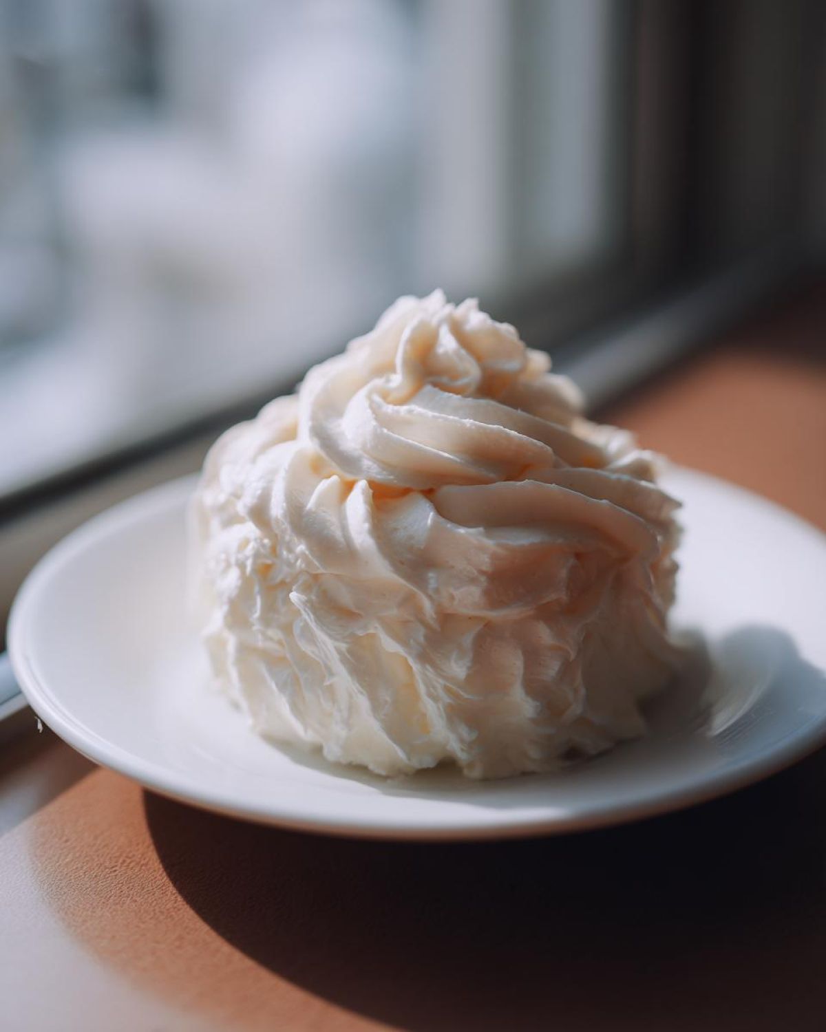 A beautiful swirl of thick, white cream cheese frosting piped onto a small white saucer, lit by natural light.