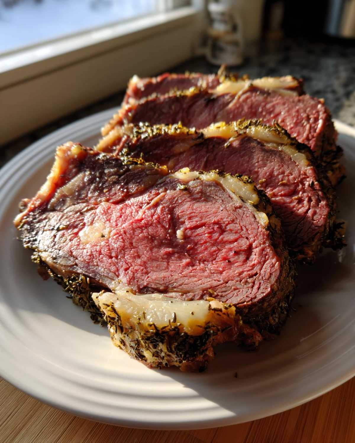 Close-up of three thick slices of medium-rare prime rib roast with a dark herb crust, resting on a white plate.
