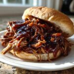 A juicy sandwich piled high with saucy pioneer woman slow cooker pulled pork on a soft bun, served on a white plate.