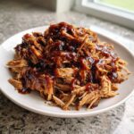 A close-up of shredded, saucy pioneer woman slow cooker pulled pork piled high on a white plate.