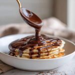 A golden spoon pours thick, glossy chocolate sauce recipe over a stack of fluffy pancakes in a white bowl.