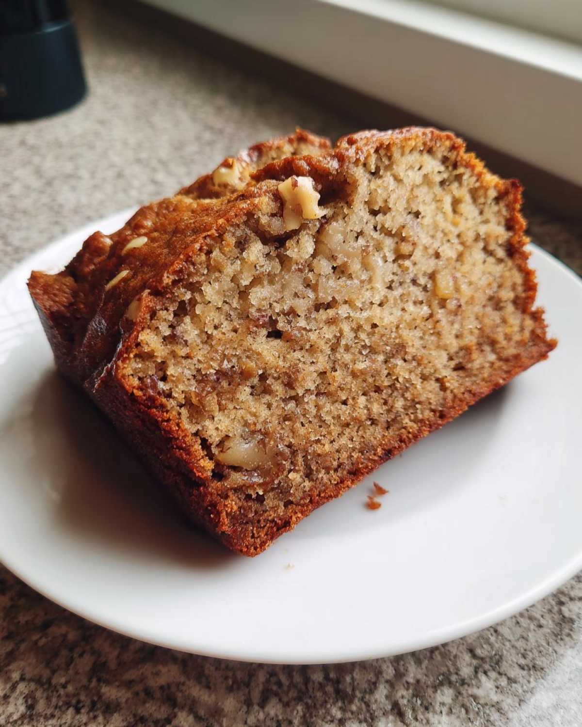 Close-up of a thick slice of moist protein banana bread showing walnuts embedded in the crumb, served on a white plate.