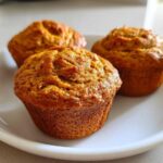 Three golden-brown protein pumpkin muffins sitting on a white plate, ready to eat.