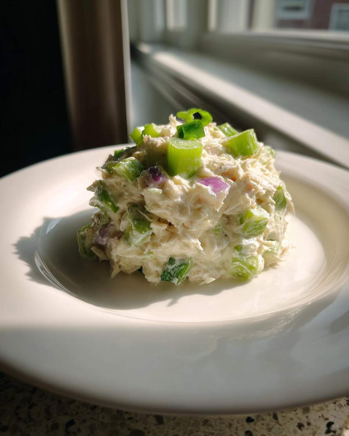 A scoop of creamy crab salad mixed with celery and topped with green onions on a white plate.