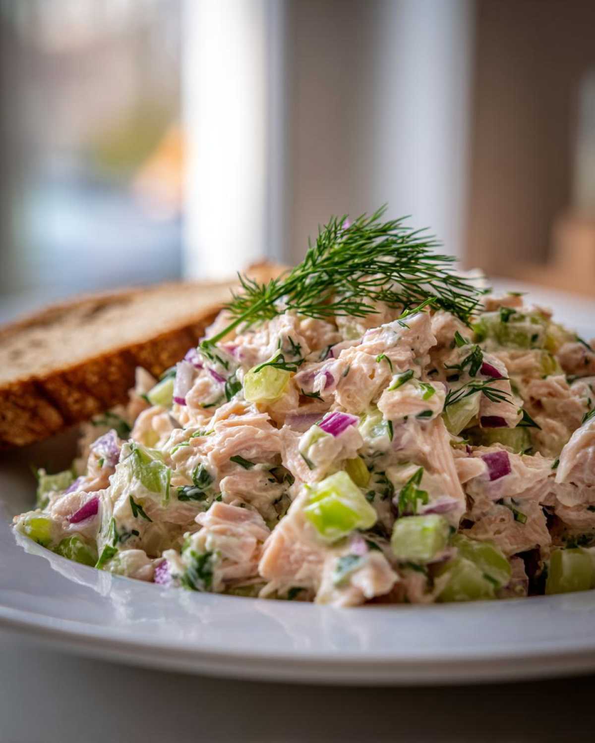 Close-up of creamy salmon salad mixed with celery and red onion, garnished with fresh dill, served next to bread.