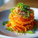 A tall stack of ribboned raw carrot salad, topped with sesame seeds and sliced green onions.