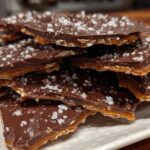 Close-up of broken pieces of saltine cracker toffee stacked on a white plate, topped with melted chocolate and flaked sea salt.