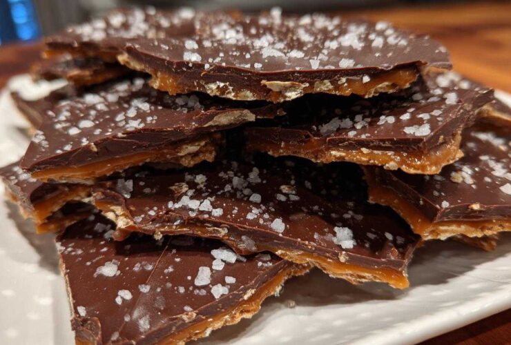Close-up of broken pieces of saltine cracker toffee stacked on a white plate, topped with melted chocolate and flaked sea salt.