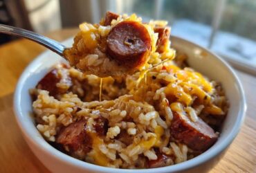 A spoonful of cheesy sausage and rice is lifted from a white bowl, showing melted cheese strings.