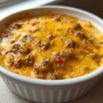 Close-up of a bubbling hot sausage cheese dip topped with melted cheddar cheese in a white ceramic dish.