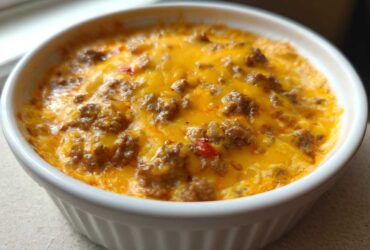 Close-up of a bubbling hot sausage cheese dip topped with melted cheddar cheese in a white ceramic dish.