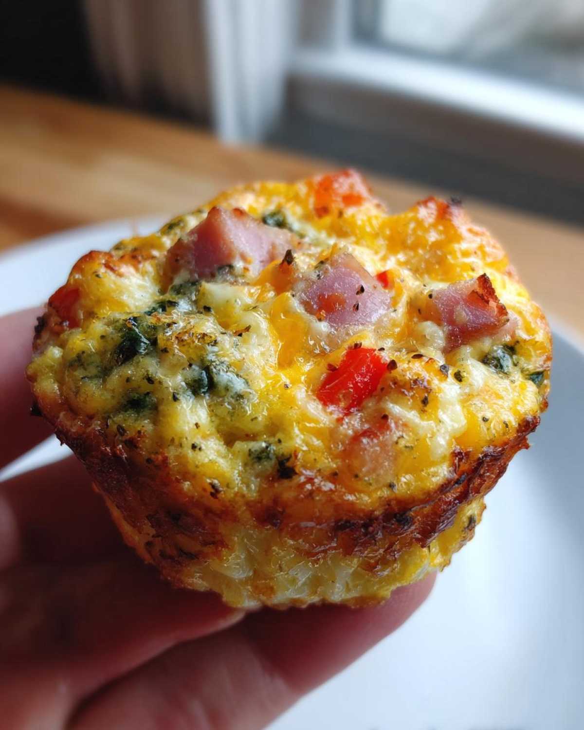 A person holding a freshly baked, savory Egg Muffin Cup topped with melted cheese, diced ham, and red peppers.
