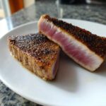 Two perfectly seared Tuna Steaks, sliced to show a rare, pink center and dark spice crust.