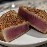 Two perfectly seared Tuna Steaks, sliced to show the rare pink center and seasoned crust.