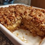 A portion removed from the baked oatmeal, showing the moist interior and crispy oat topping in a white baking dish.