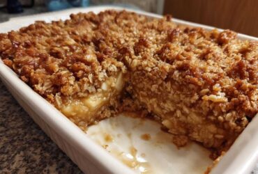 A portion removed from the baked oatmeal, showing the moist interior and crispy oat topping in a white baking dish.