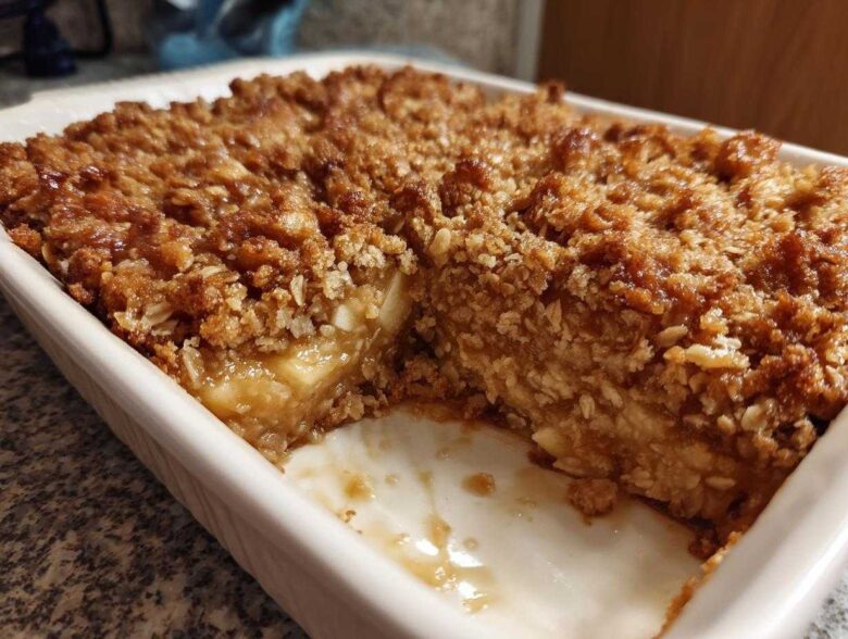A portion removed from the baked oatmeal, showing the moist interior and crispy oat topping in a white baking dish.