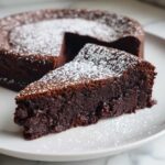 A decadent slice of flourless chocolate torte dusted heavily with powdered sugar, served on a white plate.