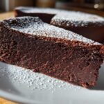 A thick slice of rich, dark flourless chocolate torte dusted with powdered sugar on a white plate.