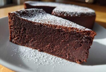 A thick slice of rich, dark flourless chocolate torte dusted with powdered sugar on a white plate.
