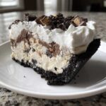 A close-up slice of a two-layer ice cream pie with a dark crust, creamy filling, whipped topping, and peanut butter cup crumbles.