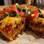 A cross-section slice of homemade Mexican pizza showing layers of seasoned beef, cheese, and topped with fresh tomatoes, olives, and green onions.