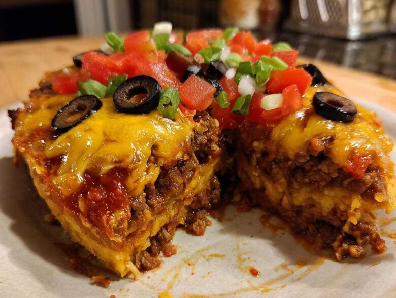 A cross-section slice of homemade Mexican pizza showing layers of seasoned beef, cheese, and topped with fresh tomatoes, olives, and green onions.
