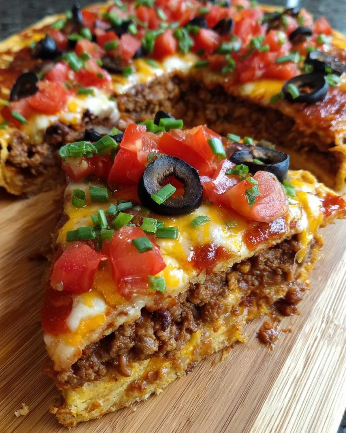 A thick slice of homemade mexican pizza showing layers of seasoned beef, melted cheese, tomatoes, olives, and green onions.