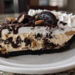 A decadent slice of two-layer ice cream pie featuring peanut butter filling, Oreo crust, whipped cream, and chocolate drizzle.