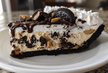 A decadent slice of two-layer ice cream pie featuring peanut butter filling, Oreo crust, whipped cream, and chocolate drizzle.