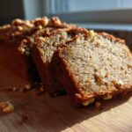 A close-up of a sliced sugar free banana bread loaf topped with walnuts, sitting on a wooden cutting board in bright sunlight.