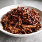 Close-up of tender, shredded Slow cooker pulled pork heavily coated in a rich, dark barbecue sauce in a white bowl.